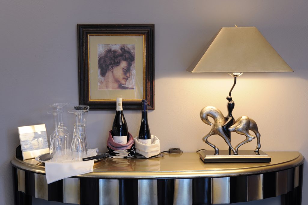Lam Lee - Begehrte Sammlerstücke Eleganter Tisch mit zwei Flaschen Wein, Gläsern, einer Lampe und einem dekorativen Pferd.