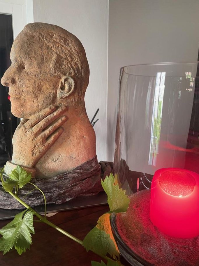 Hannelore Hilgert Eine Büste mit angeschlossener Pflanze und einem roten Kerzenlicht im Hintergrund.