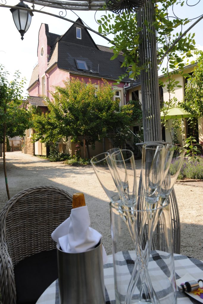 Begrüßung im Hof - Herzlich willkommen! Ein Tisch mit Gläsern und einem Sektempfang, im Hintergrund ein pinkes Haus und Grün.