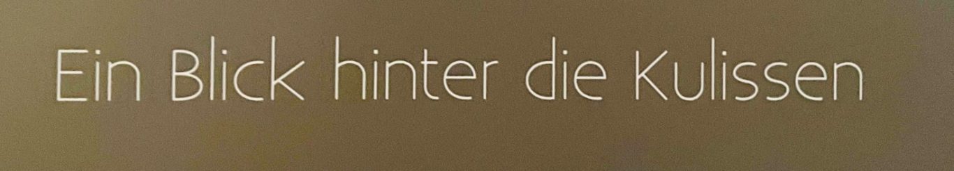 Alles was gefällt zum Mitnehmen Text: "Ein Blick hinter die Kulissen" in goldener Schrift auf braunem Hintergrund.